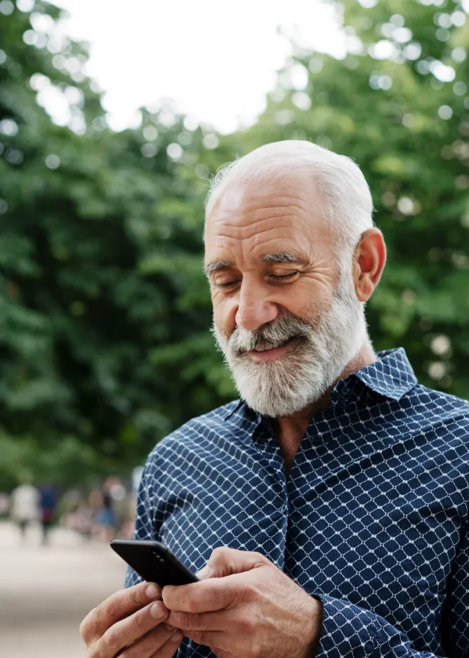 An elderly man — outdoors and smiling looking at his phone